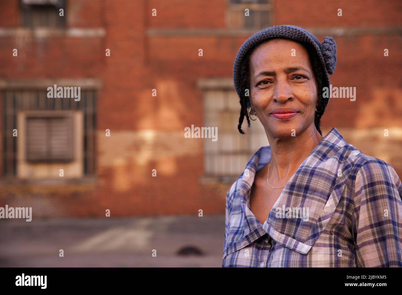 Porträt einer älteren Frau mit Strickmütze, die lächelnd in einer Gasse steht und die Kamera anschaut, Backsteinmauer im Hintergrund Stockfoto