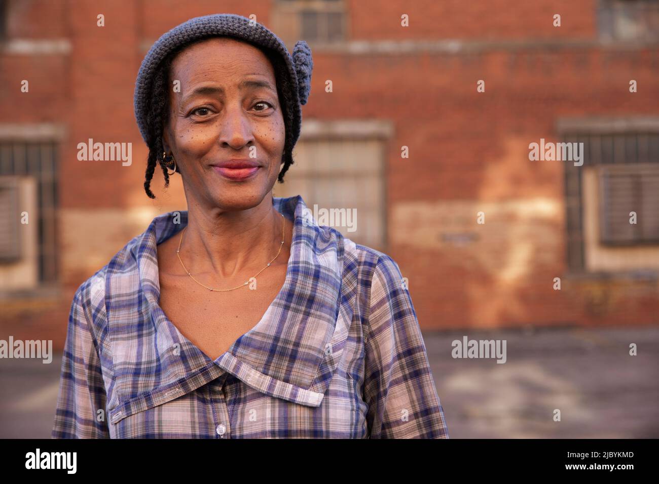 Porträt einer älteren Frau mit Strickmütze, die lächelnd in einer Gasse steht und die Kamera anschaut, Backsteinmauer im Hintergrund Stockfoto