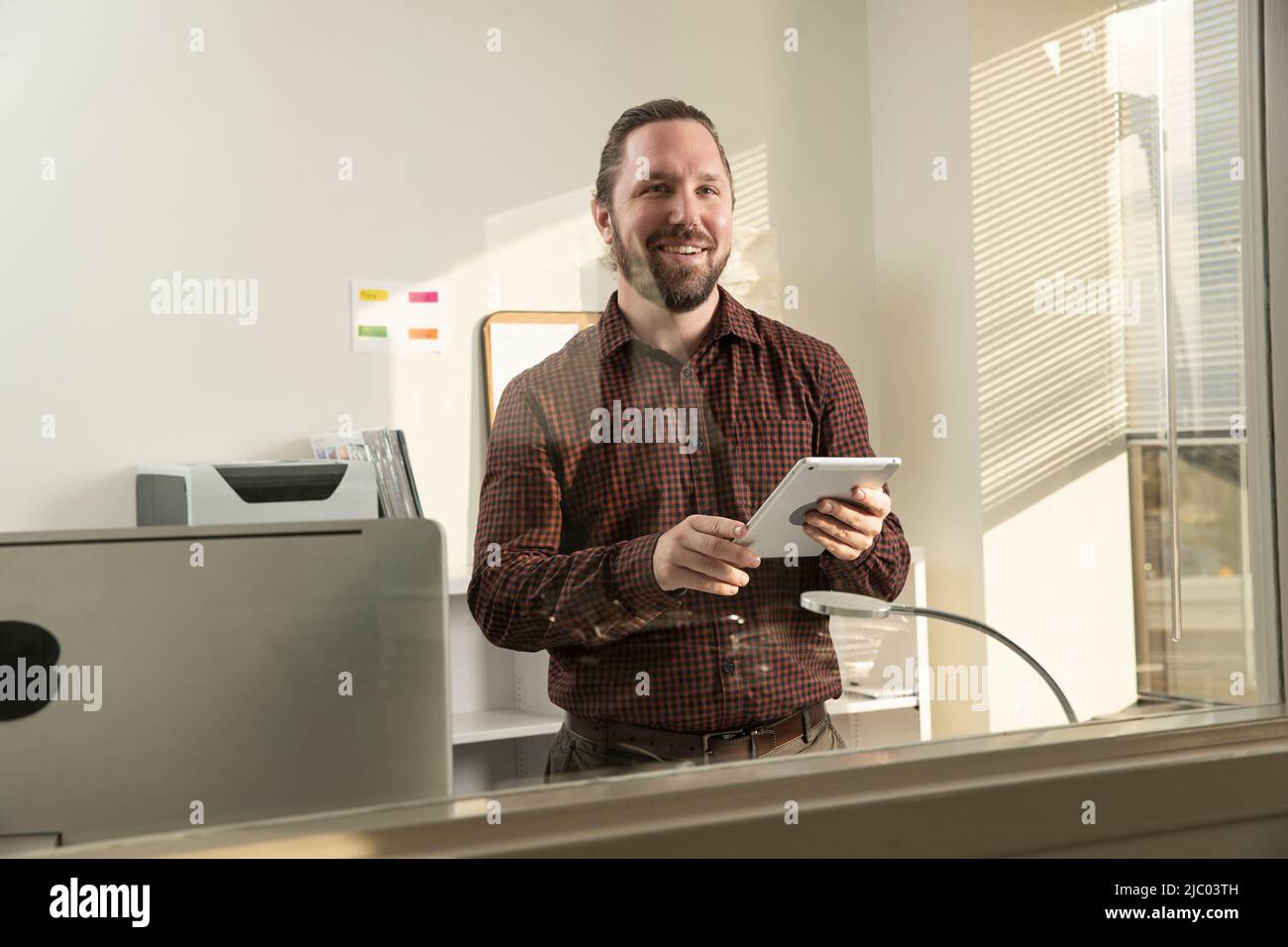 Der junge Mann blickt aus dem Fenster seines Büros und hält ein Tablet. Stockfoto