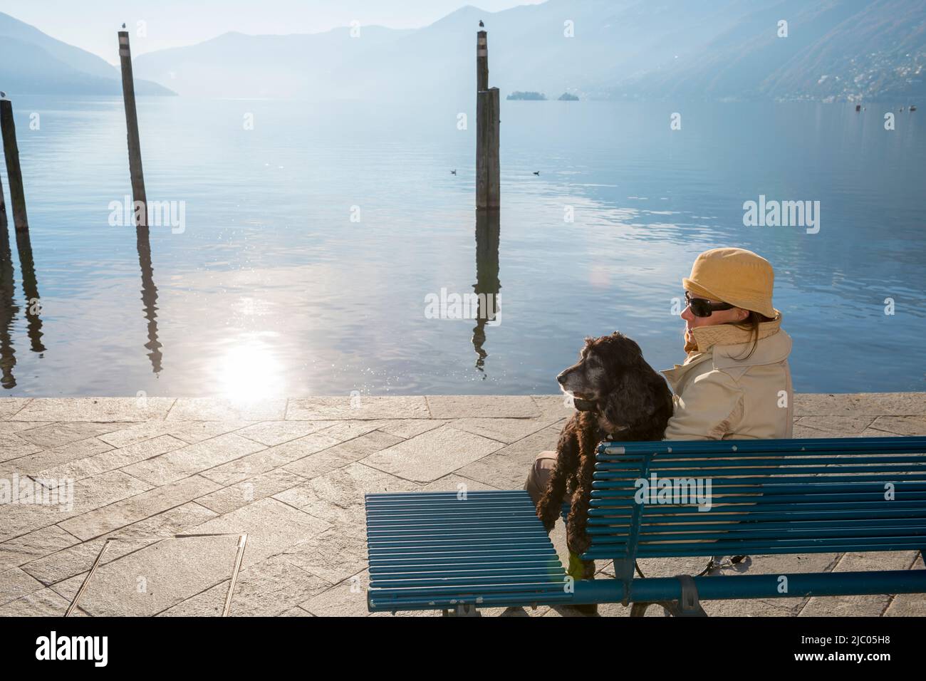 Frau sitzt auf einer Bank mit ihrem Hund an der Waterfront am Alpensee Maggiore mit Berg und Sonnenlicht in Ascona, Schweiz. Stockfoto