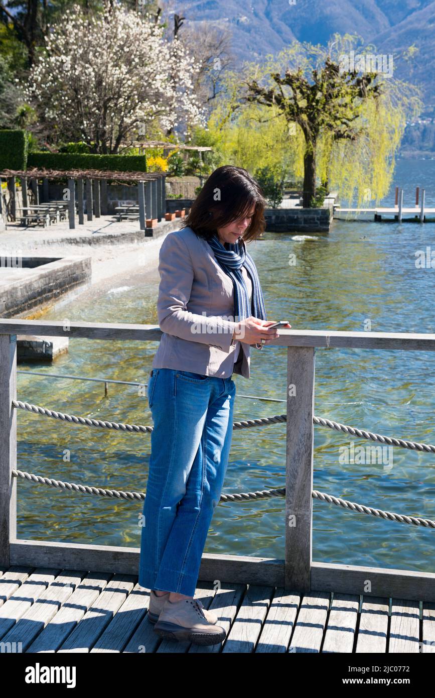 Frau steht und schief auf einem Geländer und per Telefon an der Waterfront zu einem Alpensee Maggiore mit Berg in Ascona, Schweiz. Stockfoto