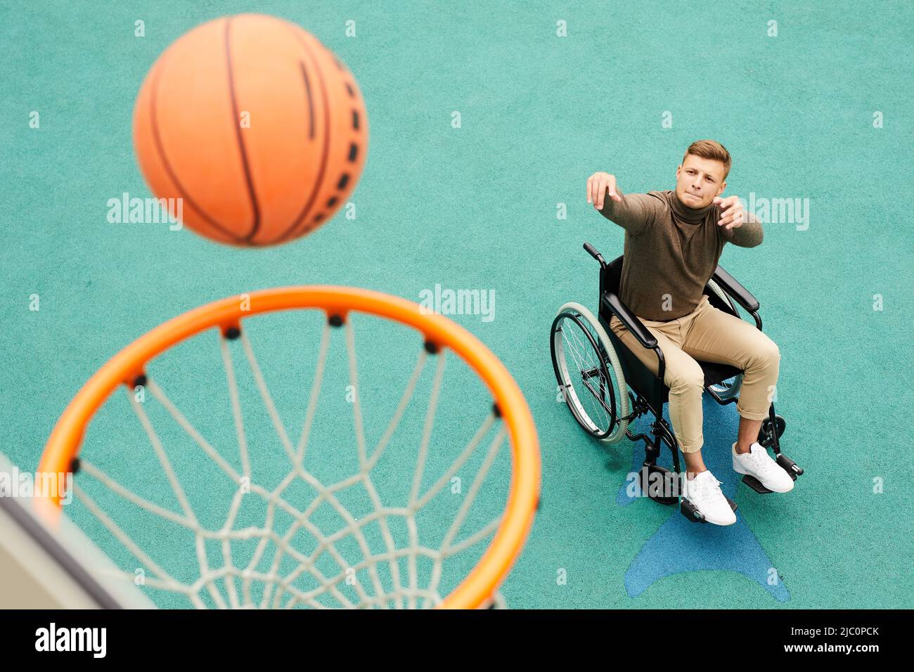 Konzentrierter junger Mann mit gelähmten Beinen, die im Rollstuhl sitzen und Lippe beißen, während er auf dem Sportplatz den Ball in den Korb wirft Stockfoto