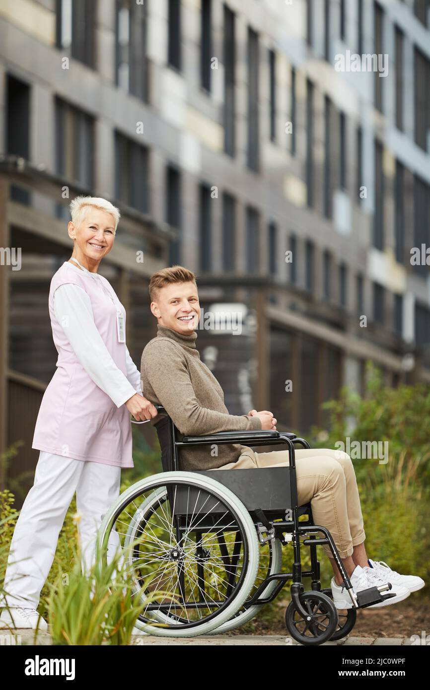 Porträt einer positiven reifen Krankenschwester in weiß und rosa Uniform hält Griffe des Rollstuhls mit behinderten Patienten, sie arbeitet mit erholt Patien Stockfoto