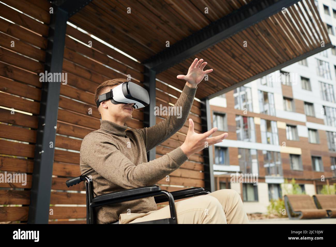 Inhalt zufriedener junger Rollstuhlfahrer in legerer Kleidung, der während der Verwendung des Simulators die Hände und die Umgebung im VR-Headset anschaute Stockfoto