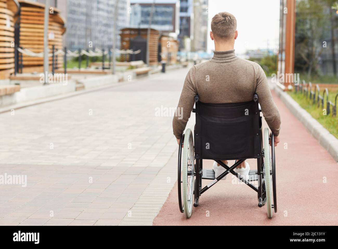 Rückansicht eines jungen Mannes mit körperlicher Behinderung, der im Rollstuhl sitzt und über die Straße der Stadt geht Stockfoto