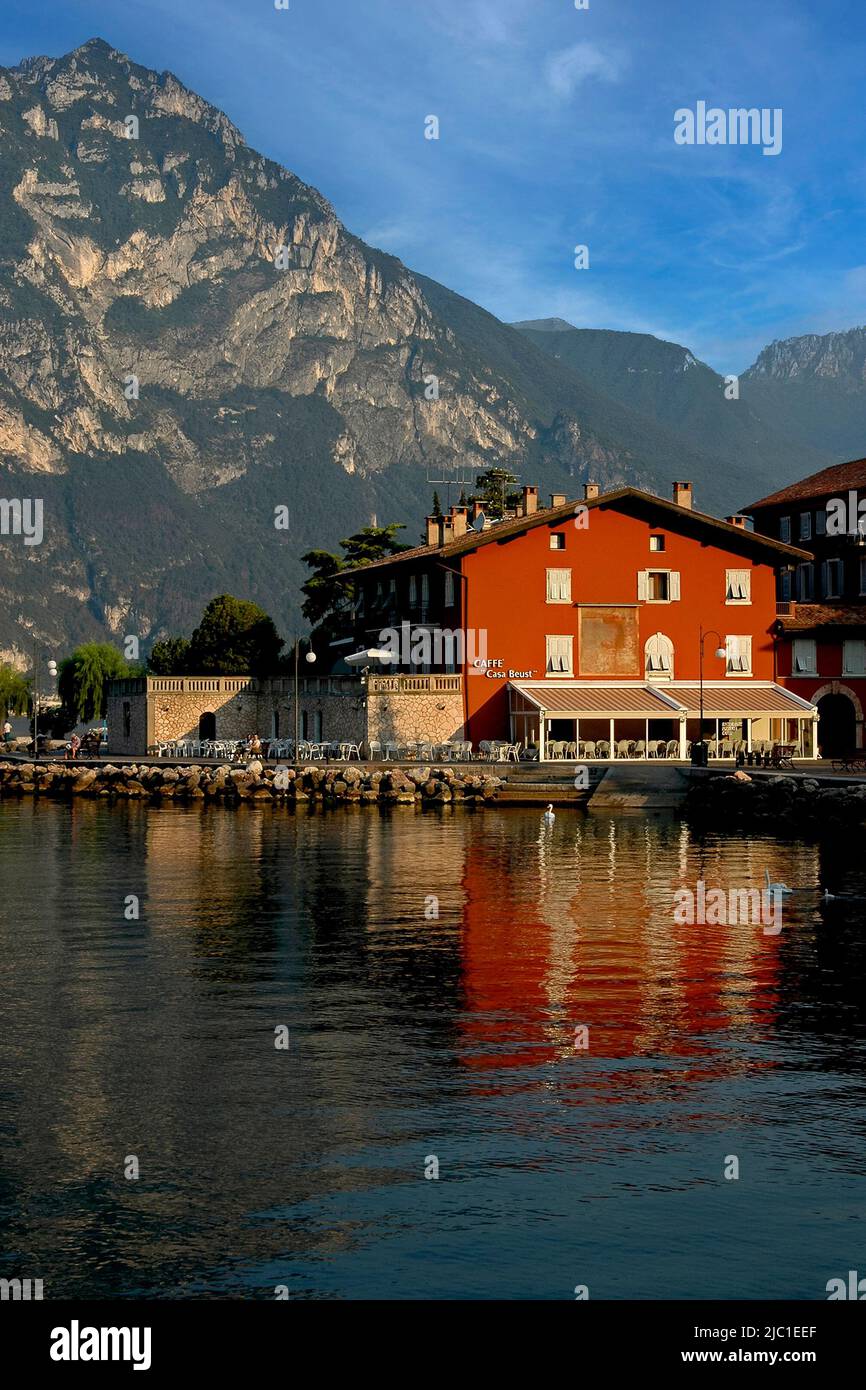 Die Casa Beust, ihre roten ockerfarbenen Wände spiegeln sich in der ruhigen Sonne des Gardasees am frühen Morgen des Julis wider, wurde 1400s als venezianische Villa am Ufer von Torbole sul Garda, Trentino-Südtirol, Italien, erbaut. Seitdem diente es als Treffpunkt für Künstler und beherbergt heute ein Restaurant, eine Pizzeria, ein Café und Apartments. Stockfoto