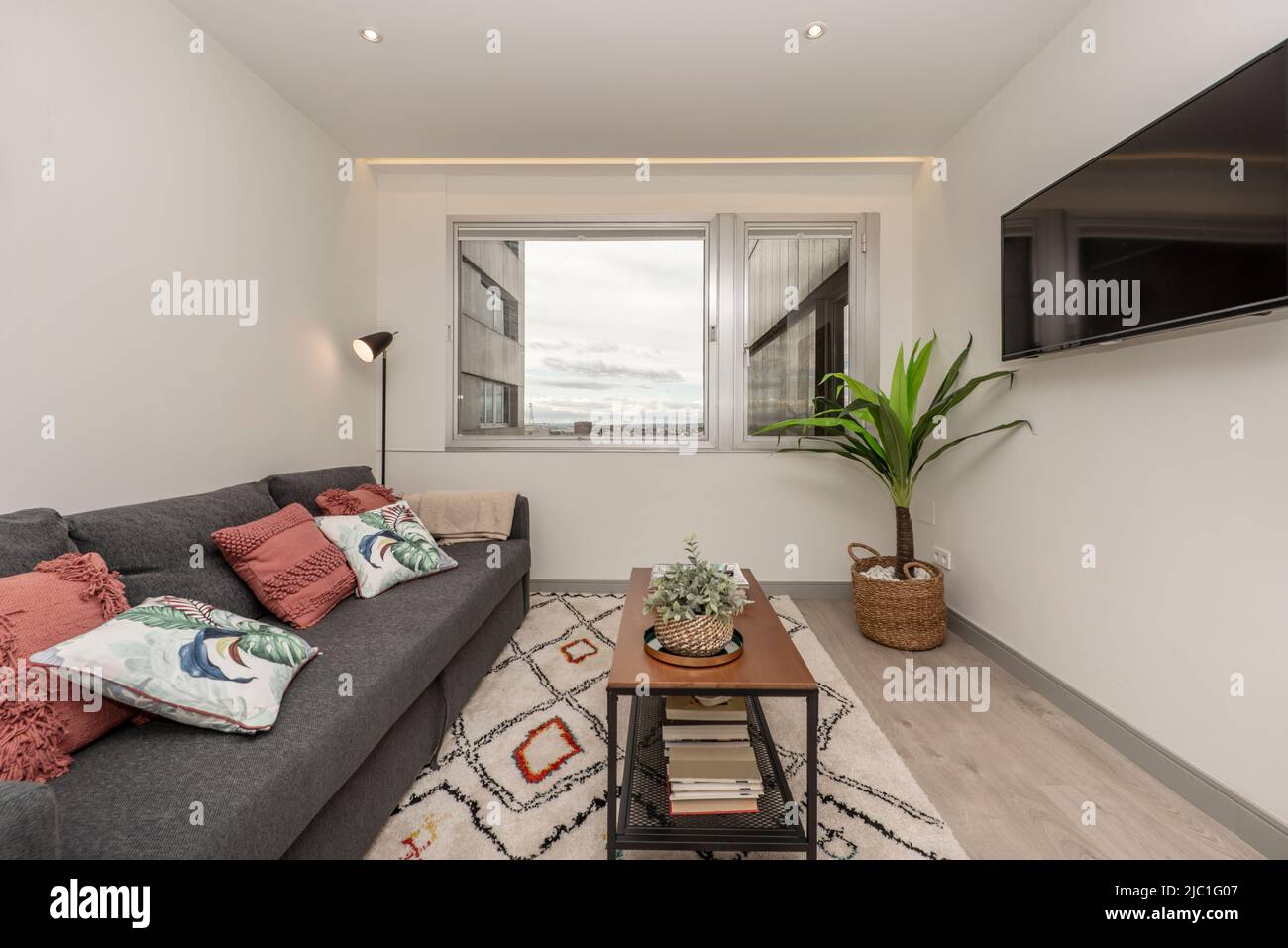 Wohnzimmer mit grauem Dreisitzer-Schlafsofa, weiß gemustertem Teppich, Beistelltisch im Industriestil mit fernseher an der Wand und Aluminiumfenster mit Aussicht Stockfoto