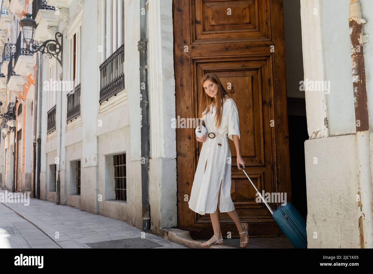 Glückliche Frau, die in valencia Handtasche und Gepäck in der Nähe der hölzernen Eingangstür hält Stockfoto