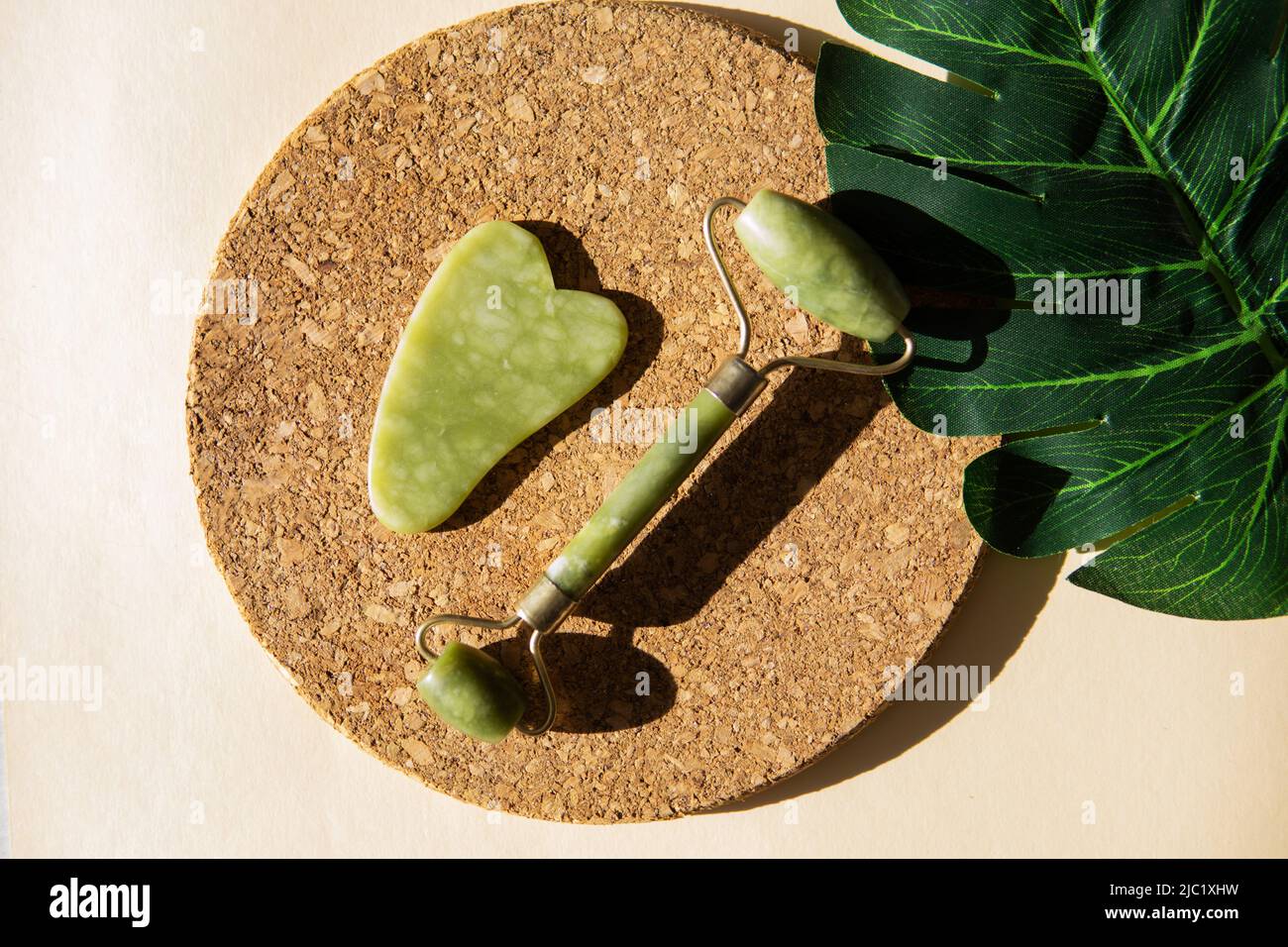 Jade Gua Sha Scraper und Face Roller Massagegerät auf einem runden Korkständer mit Monstera-Blatt. Hartes Licht, Schatten, das Konzept der Selbstversorgung. Gesichtspflege. Zer Stockfoto