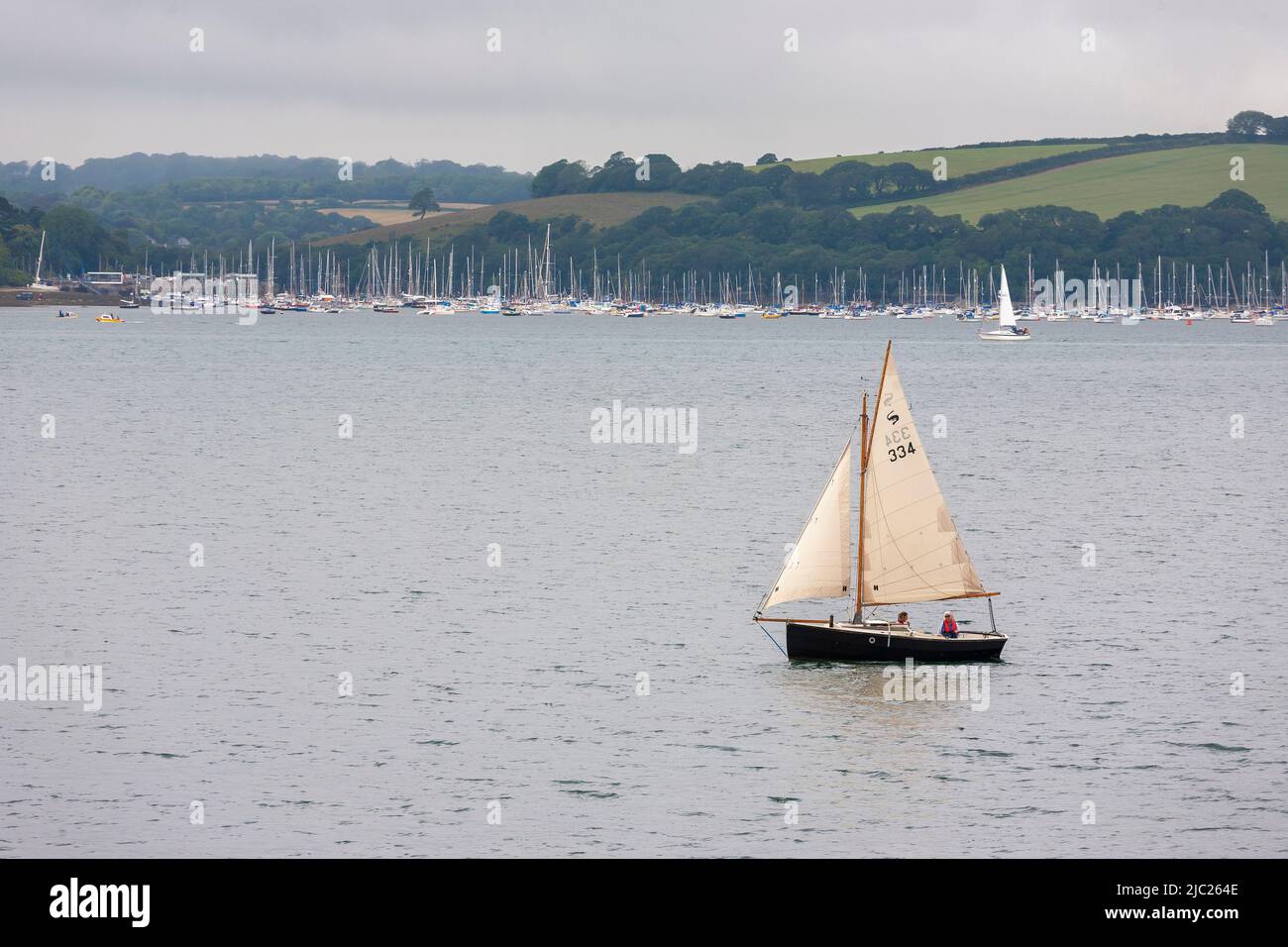 Cornish Shrimper segelt auf den Carrick Roads, mit den ausgedehnten Anlegestellen vor Mylor Creek dahinter: River Fal, Cornwall, UK Stockfoto