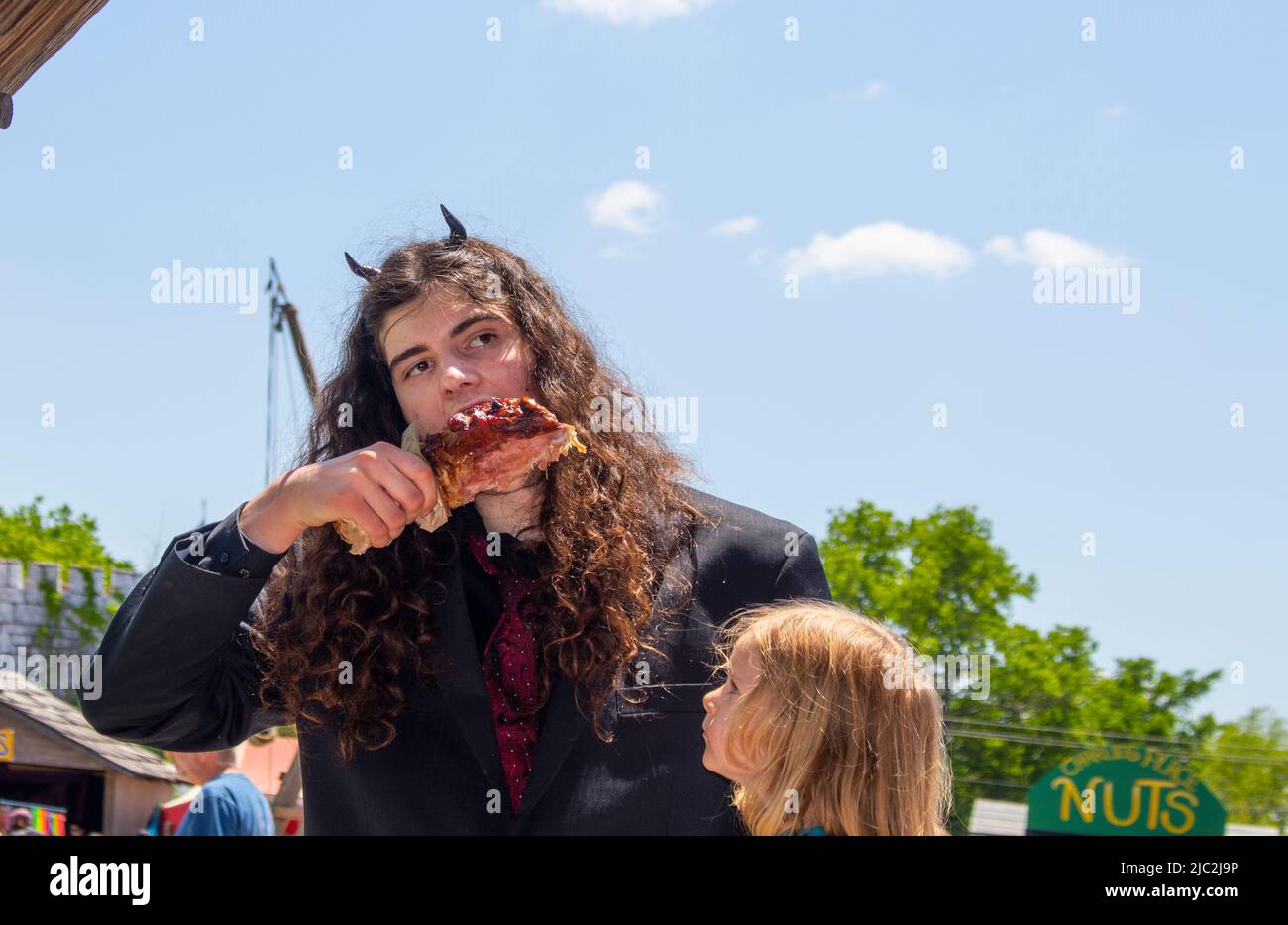 Ein junger Mann mit langen lockigen Haaren und Hörnern beißt in ein truthahn-Bein, während sein blondes Kind in Erwartung des Oklahoma Renaissance Festivals anschaut Stockfoto
