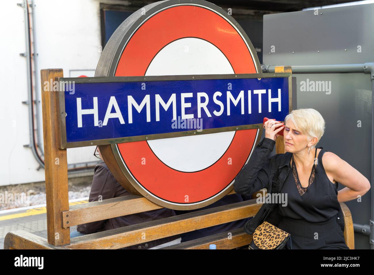 Weibliche Passagiere in schwarzem Kleid am Telefon sitzt neben Hammersmith Rundschild London England UK Stockfoto