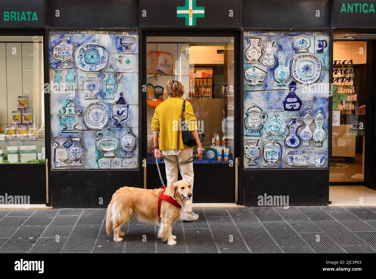 Eine Dame mit einem Hund an der Leine vor einer historischen Apotheke mit Keramikfliesen in der Nähe von San Vincenzo, Genua, Ligurien, Italien Stockfoto