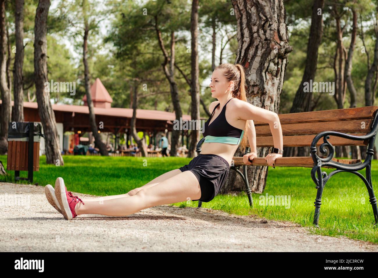 Nette kaukasische Frau, die im Stadtpark sportliche Kleidung trägt, sich im Freien auf ihr Training konzentriert, sich auf eine Parkbank lehnt und Trizeps trainiert. Stockfoto