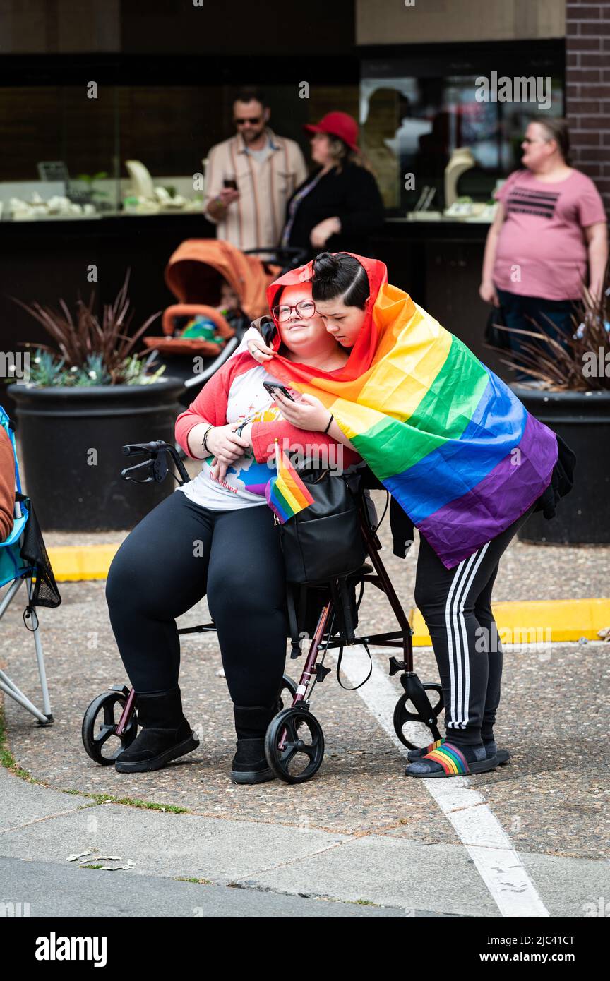 Eine junge Person trägt eine Regenbogenflagge, die eine Person auf einem Geher bei der Sonoma County Pride Parade während einer Regenschauerparty zu Beginn der Veranstaltung umarmt. Stockfoto