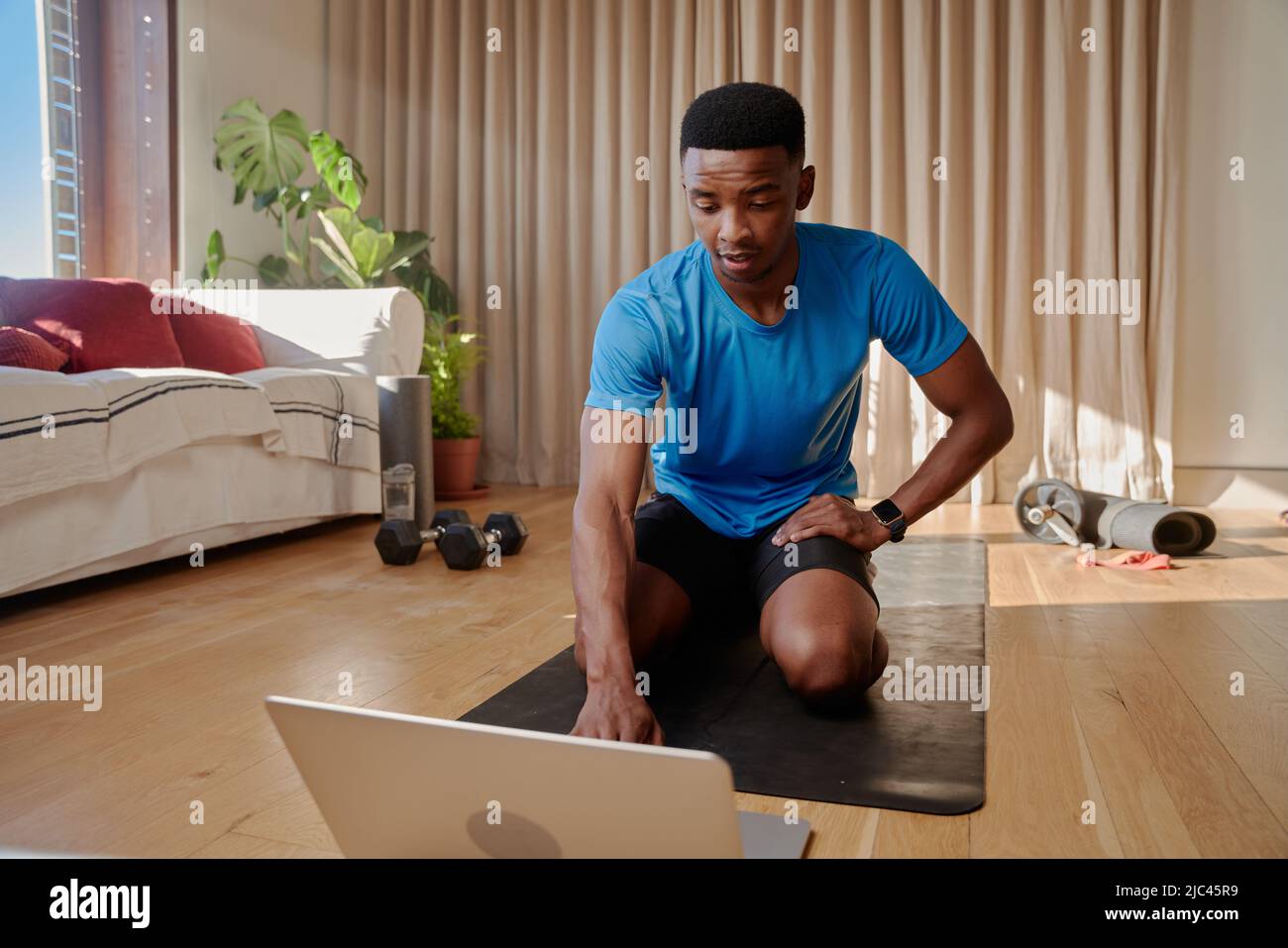 Junger afroamerikanischer schwarzer Mann, der gerade dabei ist, mit seinem Laptop an einem Online-Trainingskurs teilzunehmen, auf einer Yogamatte zu sitzen und von zu Hause aus zu trainieren Stockfoto