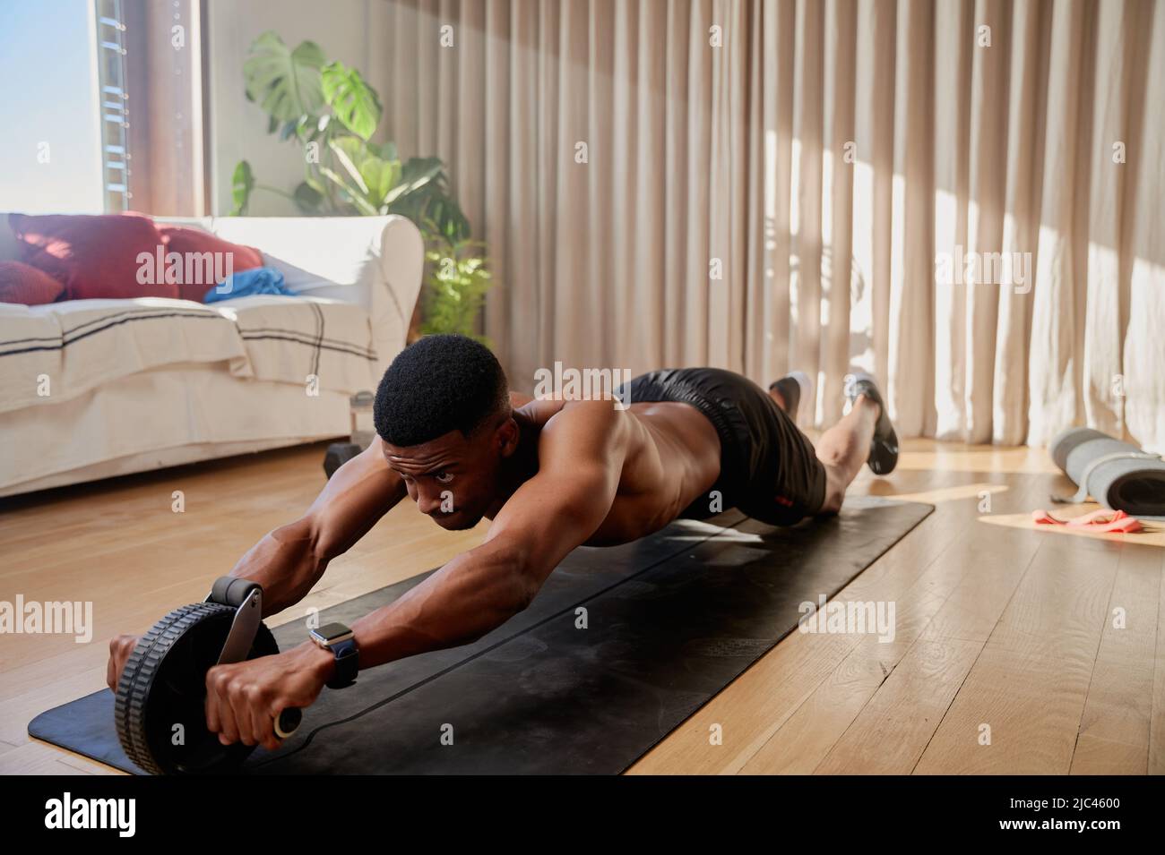 Junger afroamerikanischer schwarzer Mann, der in seinem Wohnzimmer auf seiner Yogamatte trainieren und mit einem Bauchrollerwagen trainieren Stockfoto