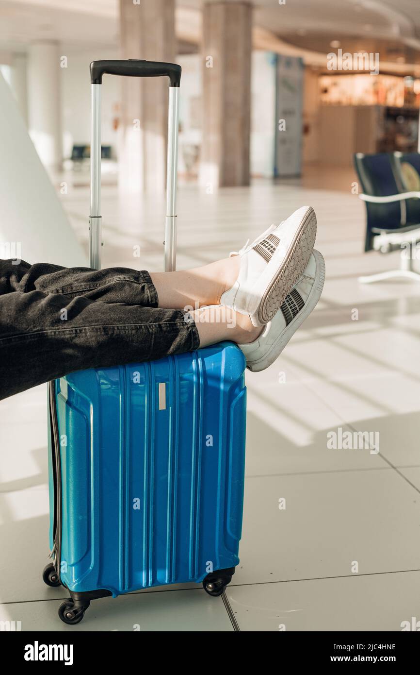 Beschnittenes Foto einer Frau in schwarzen Jeans, weißen Sneakers, gekreuzten Beinen auf türkisfarbenem Koffer im Flughafen. Stockfoto