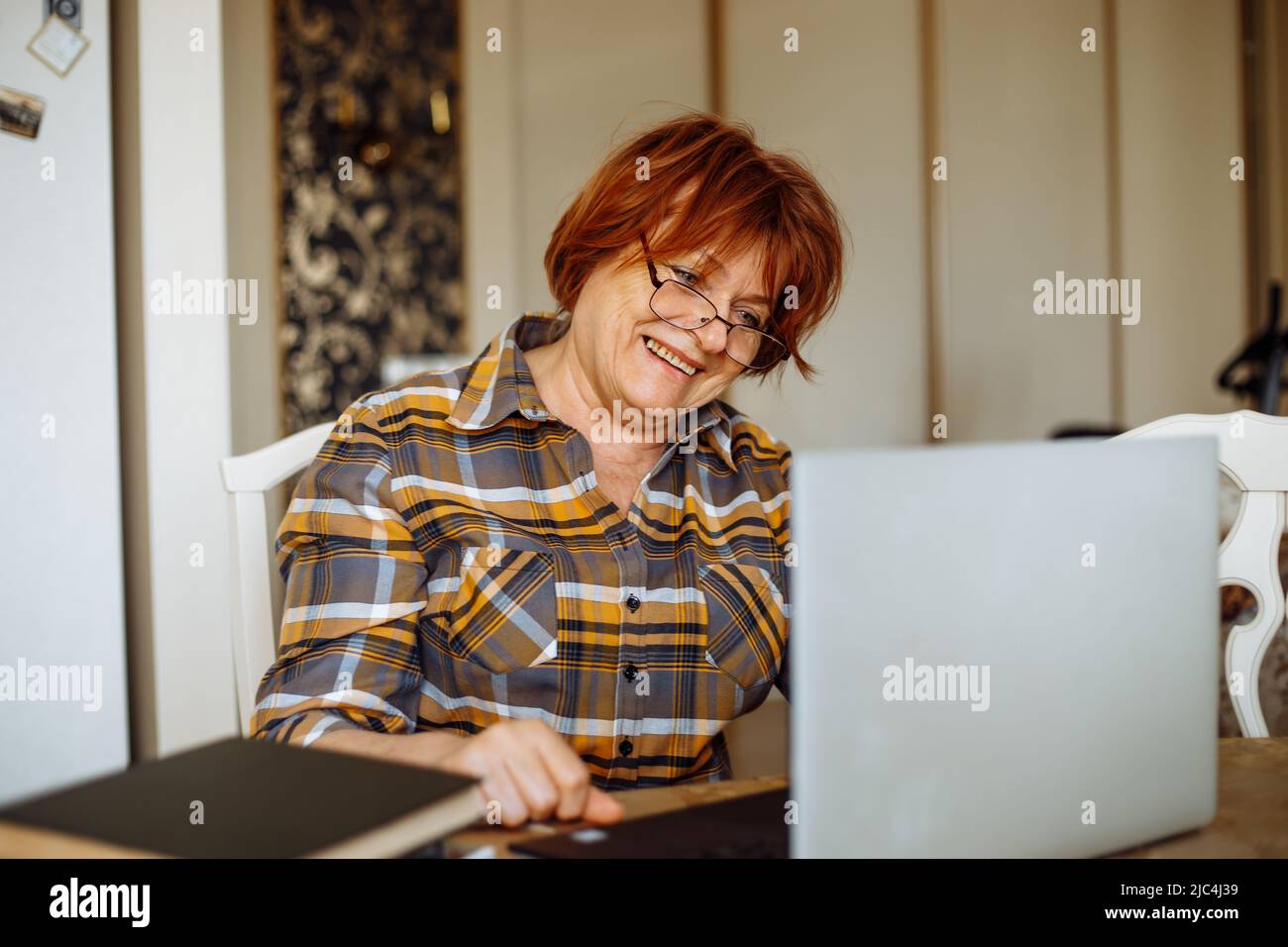 Konzentrierte aktive ältere Frau in einer Brille mit Laptop, die für ein Online-Studium in der Küche tippt. Surfen Sie im Internet, chatten Sie Stockfoto