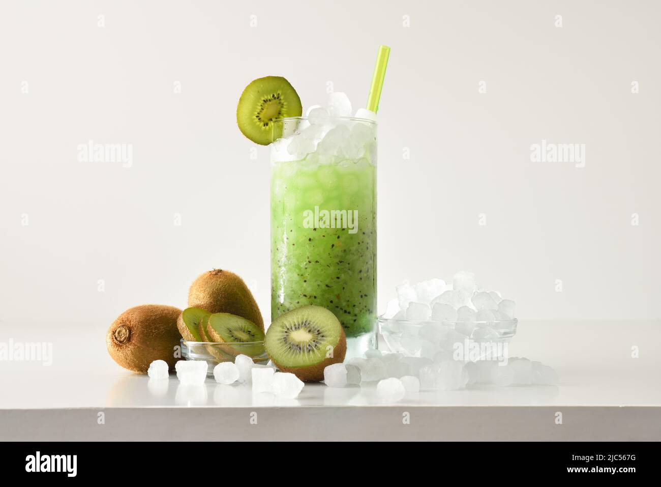 Kiwi-Slush-Getränk in hohem Glas mit Früchten und zerkleinerten Eis um ihn herum auf weißem Tisch mit isoliertem Hintergrund. Vorderansicht. Horizontale Zusammensetzung. Stockfoto