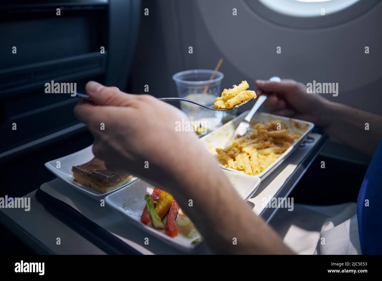 Passagier isst Flugzeugeessen mit Metallbesteck. Menü mit Pasta, Gemüse, Dessert und Getränken auf dem Tablett während des Langstreckenfluges. Stockfoto