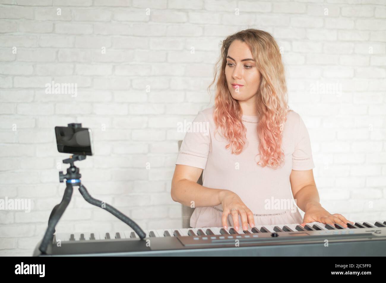 Eine Frau spielt das elektronische Klavier und führt einen Videoblog auf einem Smartphone durch. Ein Mädchen nimmt Videos von einem Telefon auf einem Stativ auf. Weibliche Musiklehrerin arbeitet Stockfoto