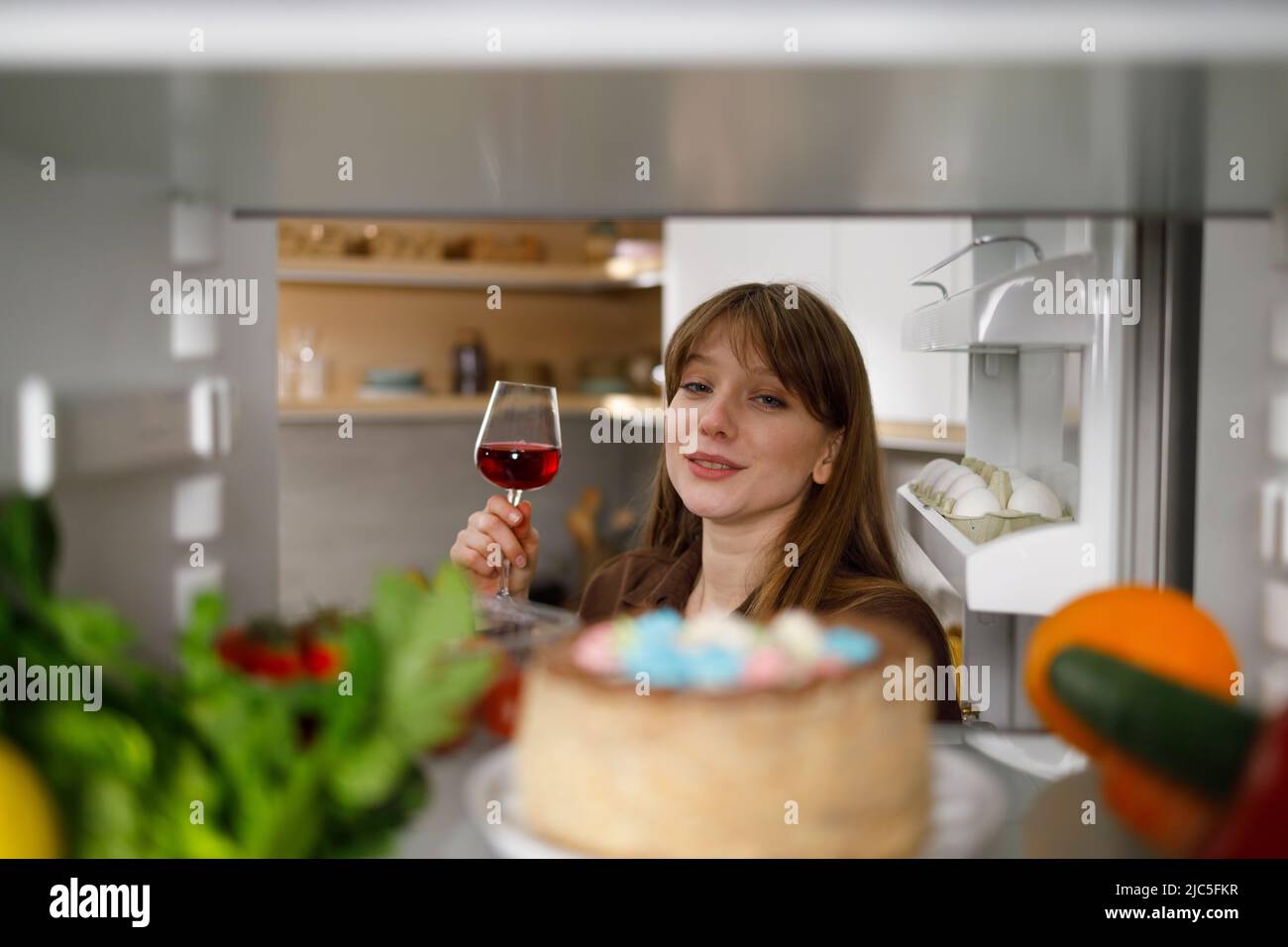 Betrunkene Frau mit einem Glas Rotwein wählt im Kühlschrank Lebensmittel aus Stockfoto