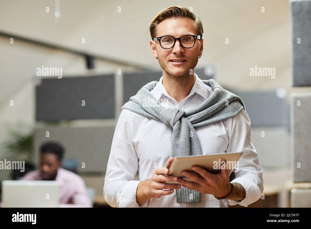 Lächelnder, stilvoller Analytiker mit Stoppeln, die im Büro der Firma SMM stehen und moderne Geräte verwenden Stockfoto
