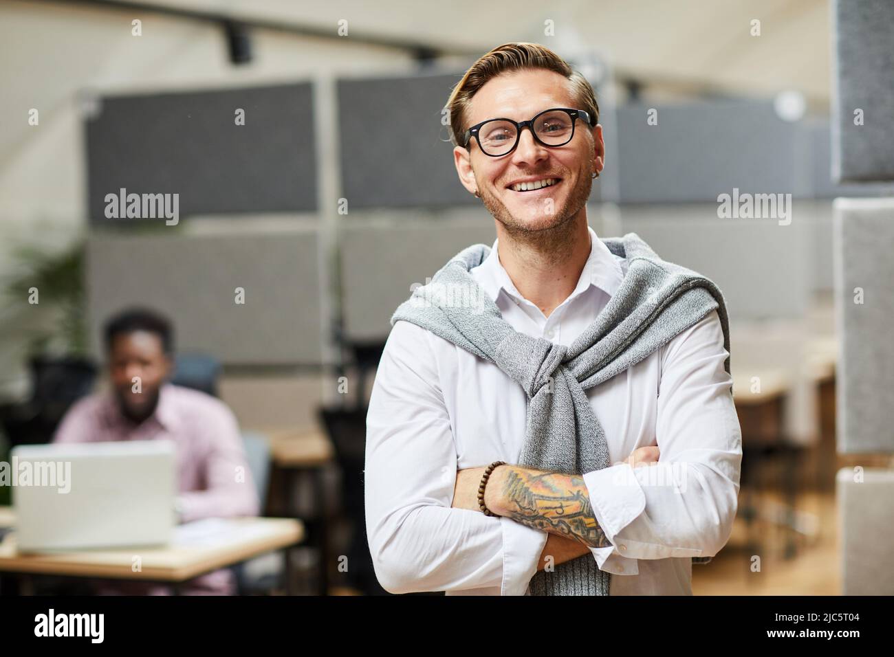 Portrait eines positiven begeisterten jungen App-Entwicklers mit Pullover um den Hals gewickelt und mit gekreuzten Armen im modernen Büro stehend Stockfoto