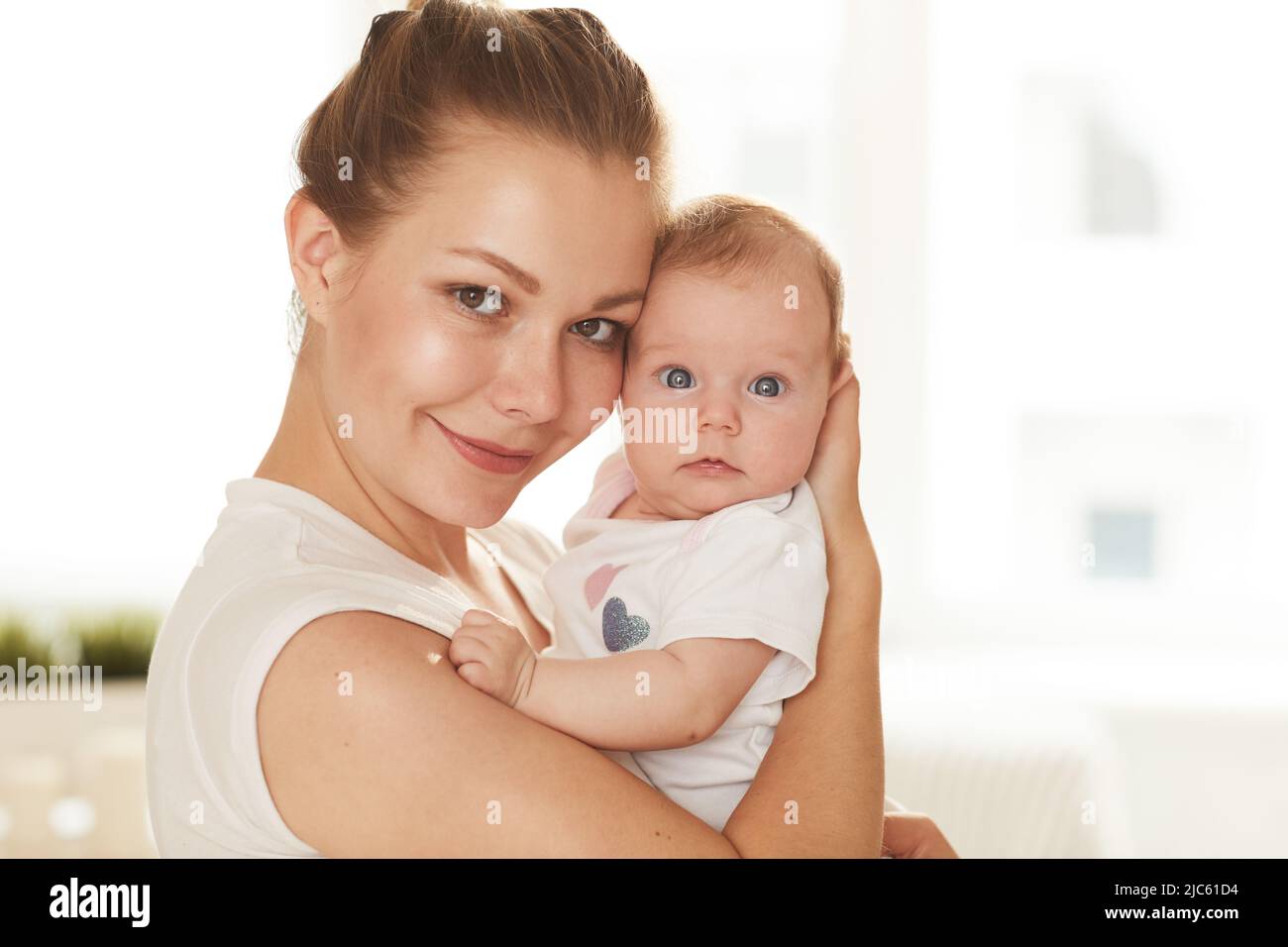 Junge lächelnde Mutter im weißen T-Shirt und ihr kleines Baby mit großen blauen Augen, die auf die Kamera schauen Stockfoto