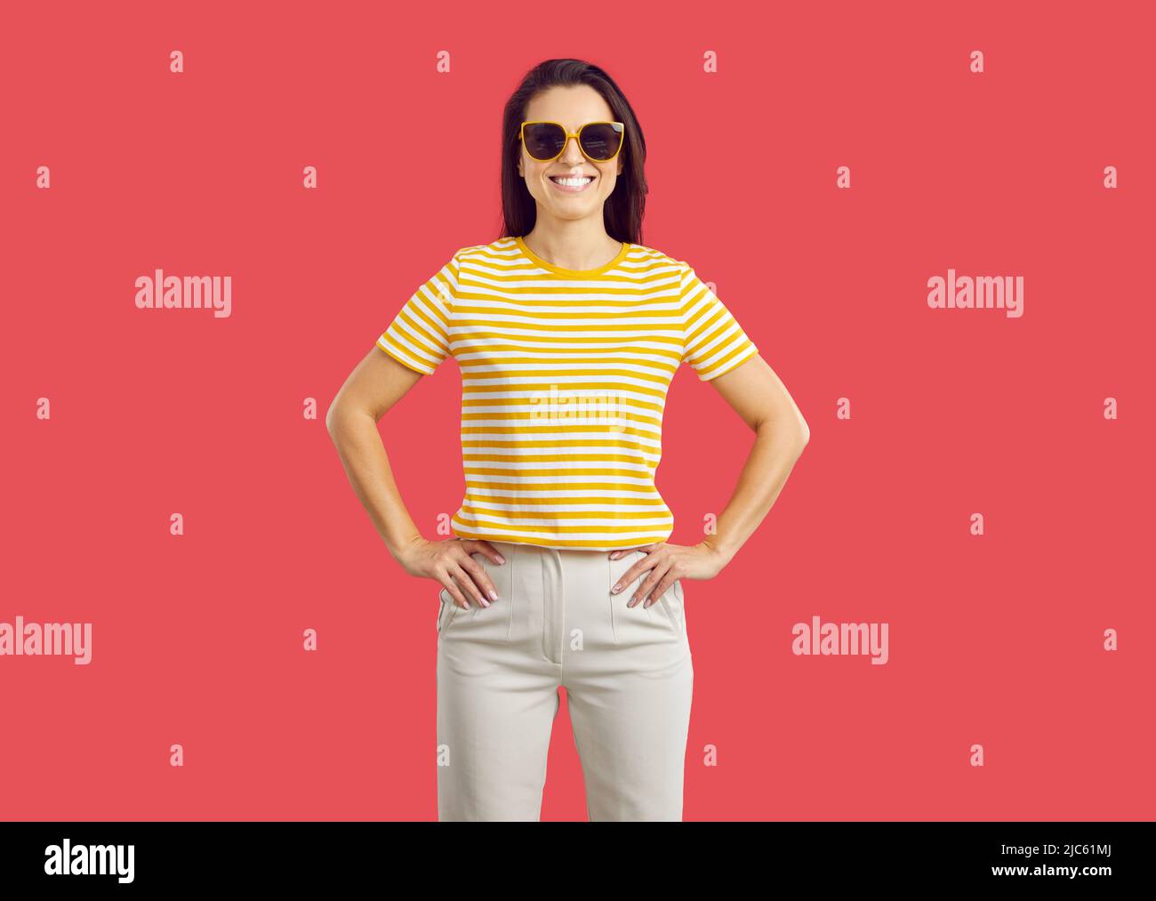 Fröhliche lächelnde Frau in lässigem Outfit und Sonnenbrille, die isoliert auf rotem Hintergrund steht Stockfoto