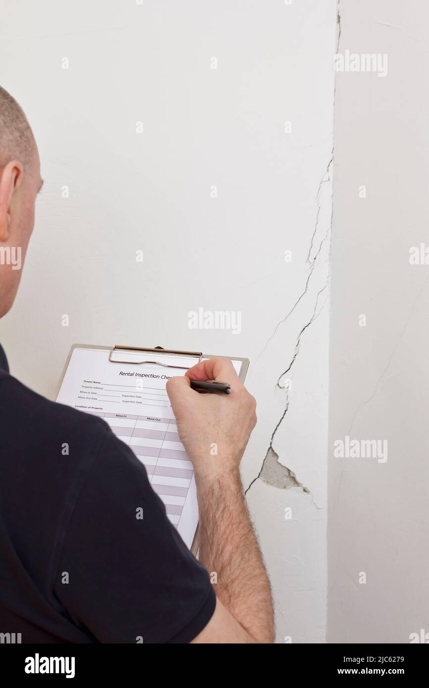 Mann mit Inspektions-Checkliste vor einer weißen Wand mit langem Riss oder Riss und fehlendem Gipsstück, Mietschaden-Konzept. Stockfoto
