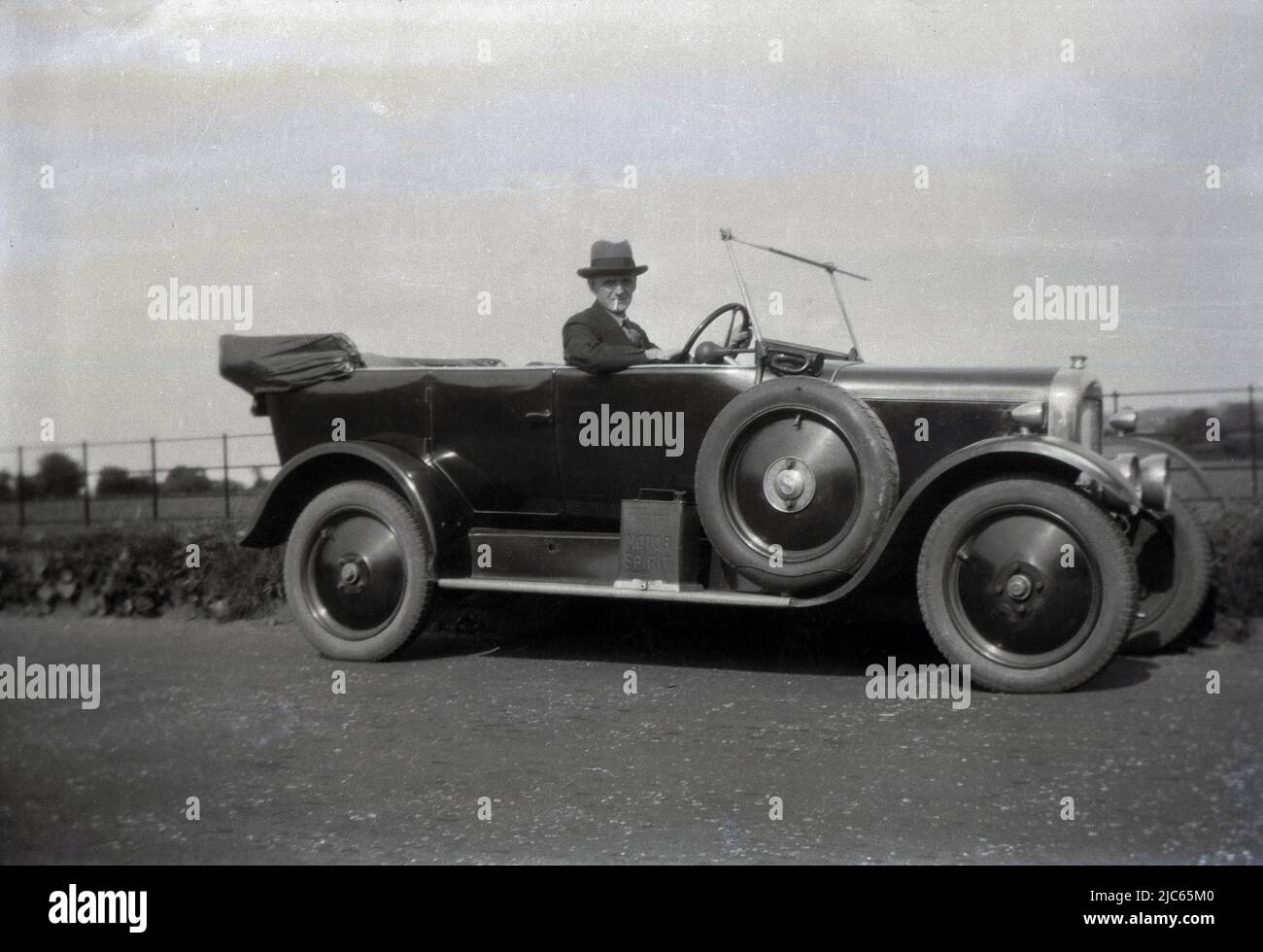 1938, historisch, ein Gentleman mit Hut und Zigarette im Mund, der für ein Foto in seinem Tourenwagen sitzt, der auf einer Landstraße in England, Großbritannien, geparkt ist. Der möglicherweise im 1920s hergestellte Wagen mit offenem Oberdeck verfügt außen neben dem Fahrer über ein einfaches Druckhorn und an der Umrandung unter der Fahrertür über eine Dose Motorgeist. Ungewöhnlich ist, dass die Räder solide sind, während die meisten Räder in dieser Zeit Speichen hatten. Das Reserverad an der Außenseite des Fahrzeugs hat den Buchstaben S in der Nabe. Stockfoto