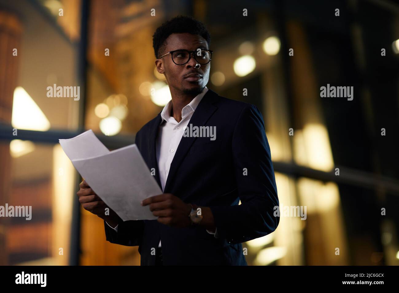 Seriös schöner afroamerikanischer Geschäftsmann mit Brille, der über die dunkle Straße läuft und die Papiere überprüft Stockfoto