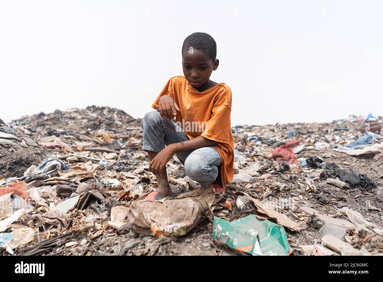 Verzweifeltes afrikanisches Kind, das auf einer Müllhalde sitzt und den Plastikmüll ansieht. Konzept der Umweltgerechtigkeit. Stockfoto