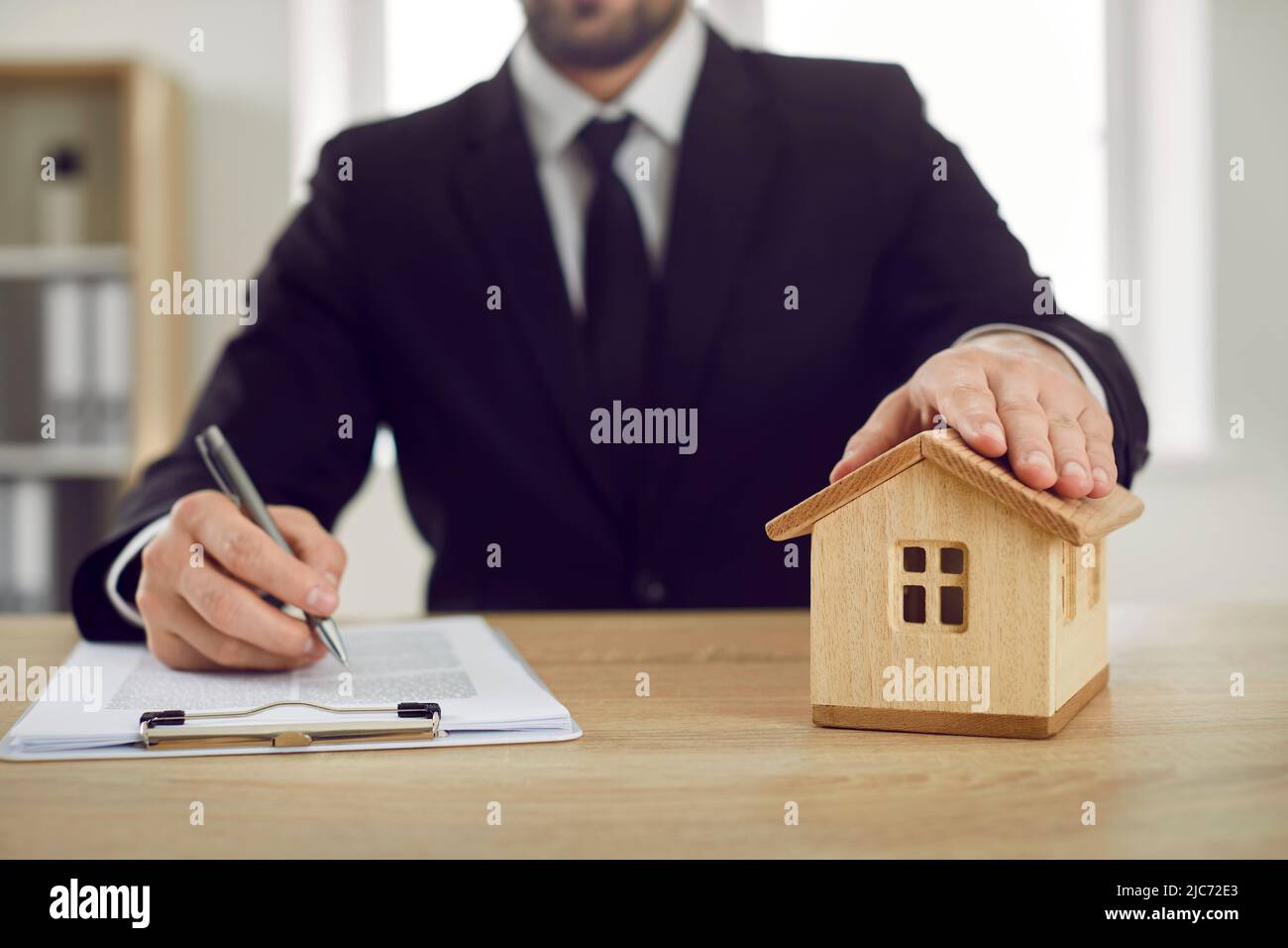 Der Mann im Büro des Immobilienmaklers legt seine Hand auf das Spielzeughaus und unterzeichnet einen Hypothekenvertrag Stockfoto