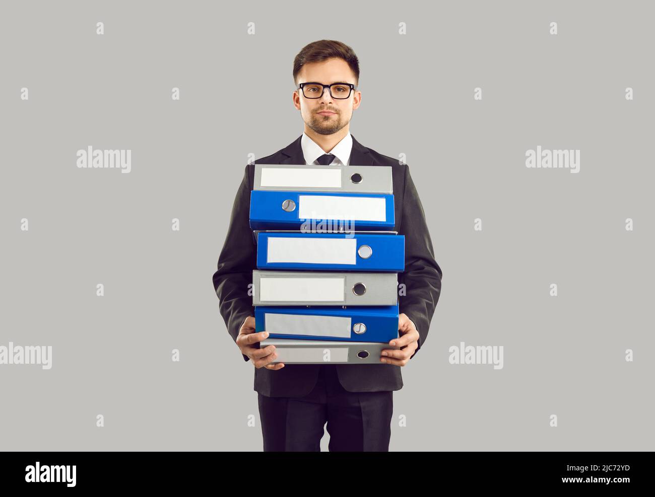 Ernsthafter männlicher Büroangestellter, der auf grauem Hintergrund einen Stapel großer Ordner für Dokumente hält. Stockfoto