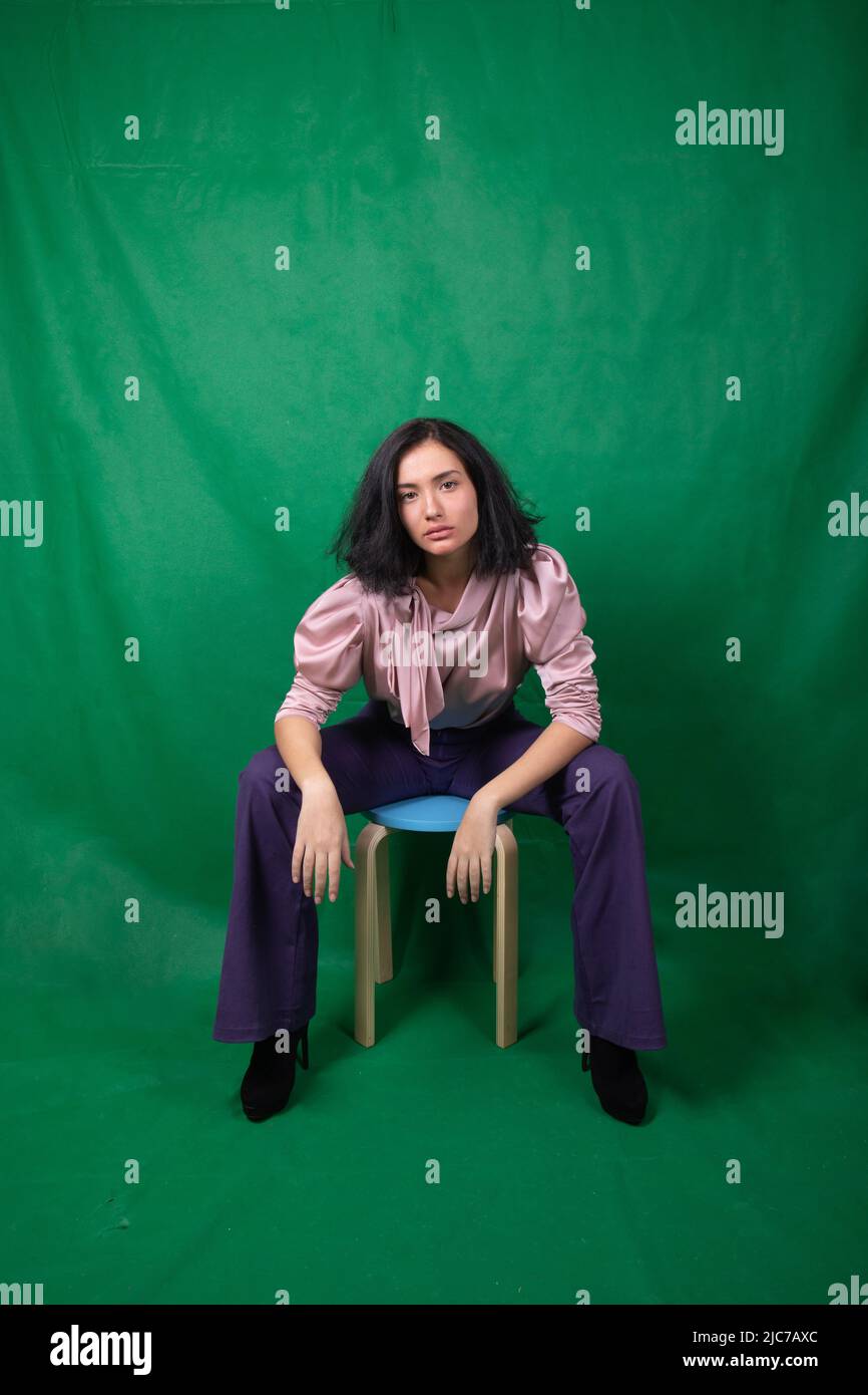 Junge hübsche Model sitzt auf einem blauen Hocker und ruht ihre Ellbogen auf den Knien im Studio Stockfoto