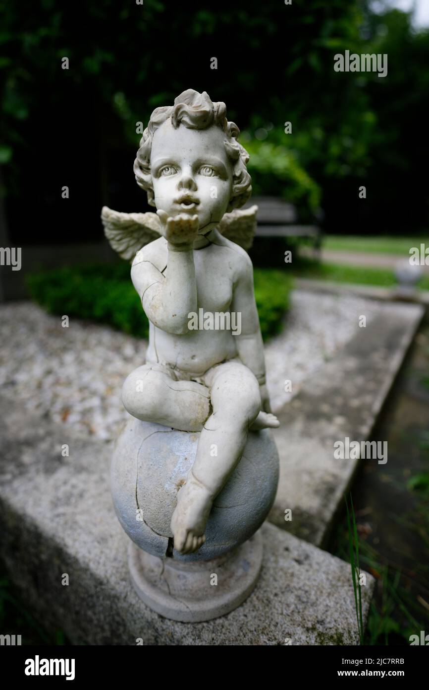 Beschädigte kleine Engelsfigur sendet Liebesgrüße, die auf einer Steinkugel an einem Grab sitzen Stockfoto