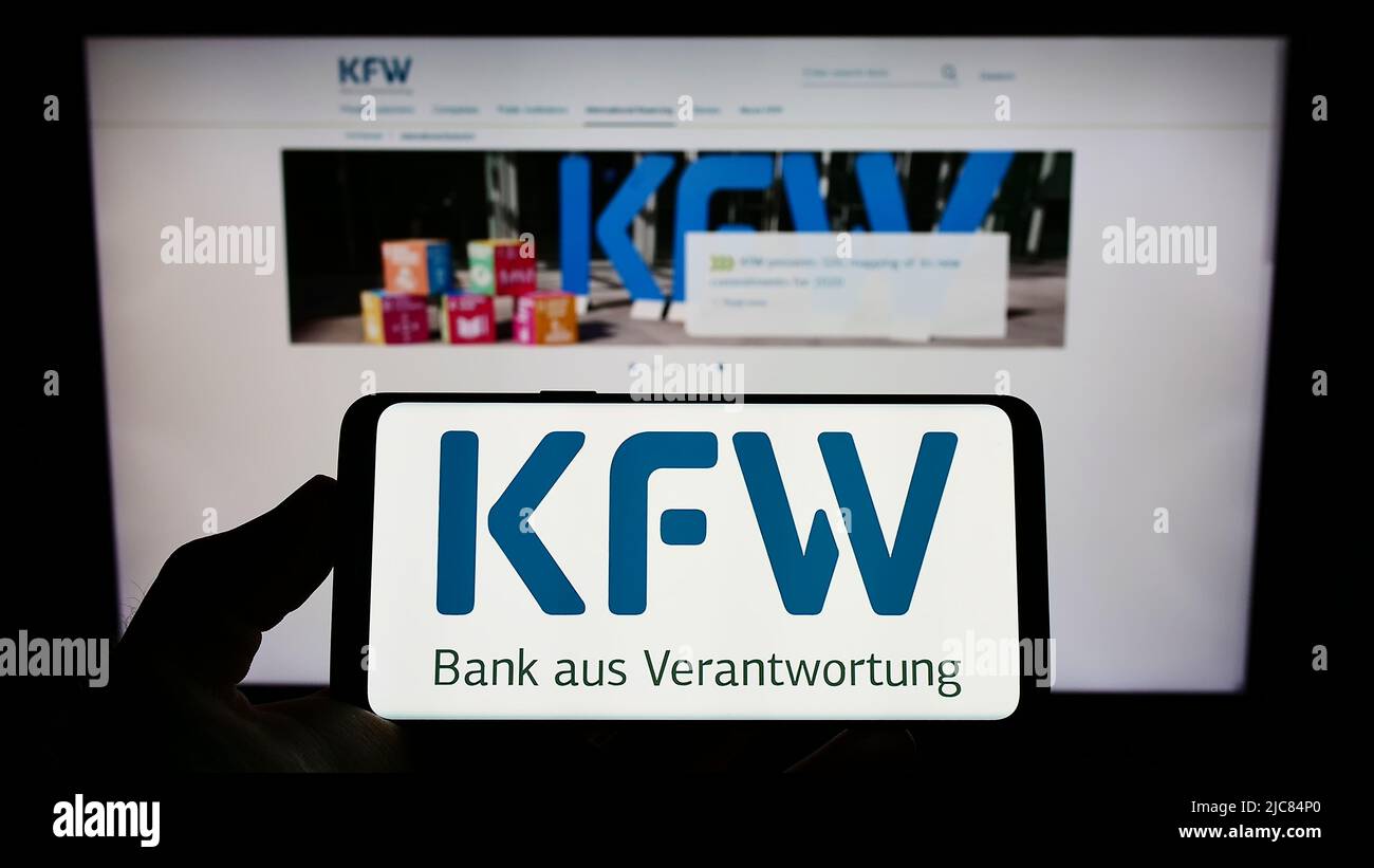 Person, die das Mobiltelefon mit dem Logo der Kreditanstalt für Wiederaufbau (KfW) auf dem Bildschirm vor der Webseite hält. Konzentrieren Sie sich auf die Telefonanzeige. Stockfoto