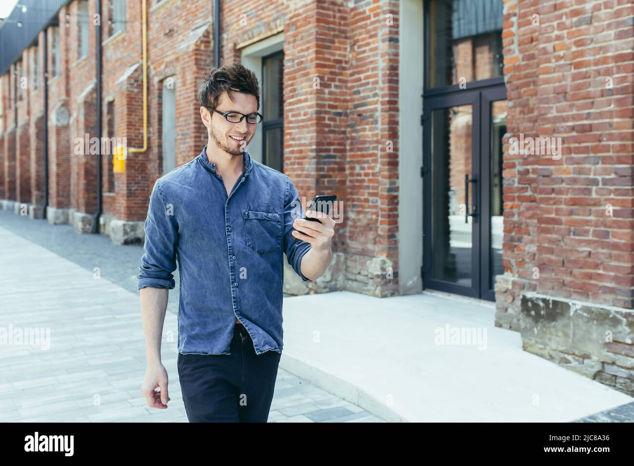 Ein junger, gutaussehender Mann mit Brille geht mit einem Mobiltelefon eine Stadtstraße entlang und sucht auf einem GPS-Navigator nach einem Platz und einer Straße. Stockfoto