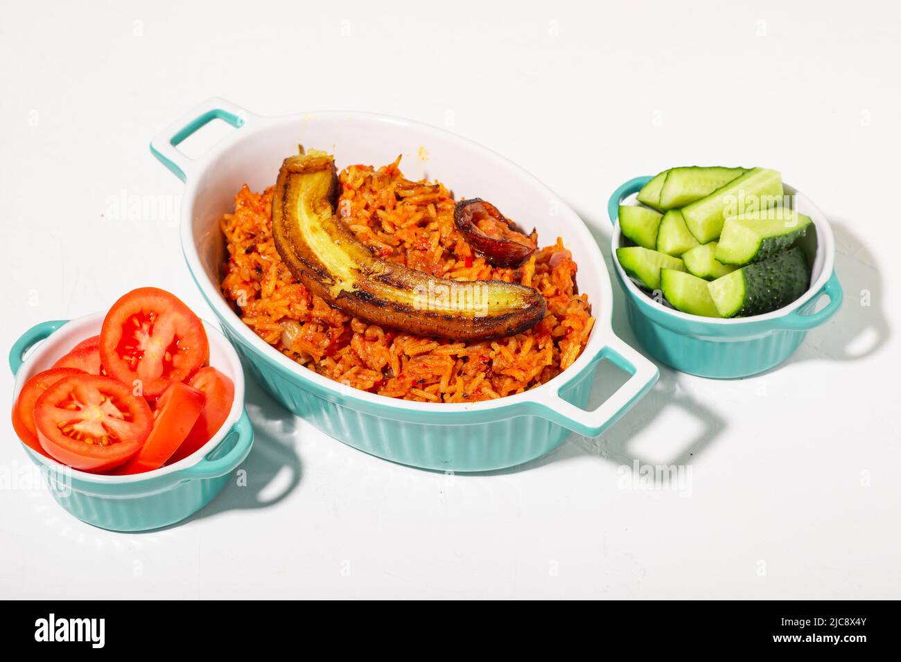 Reisjollof mit gebratener Banane. Frisches Gemüse - Tomaten und Gurken. Weißer Hintergrund. Traditionelle nigrische Reisgerichte mit Tomaten, Zwiebeln und Spic Stockfoto