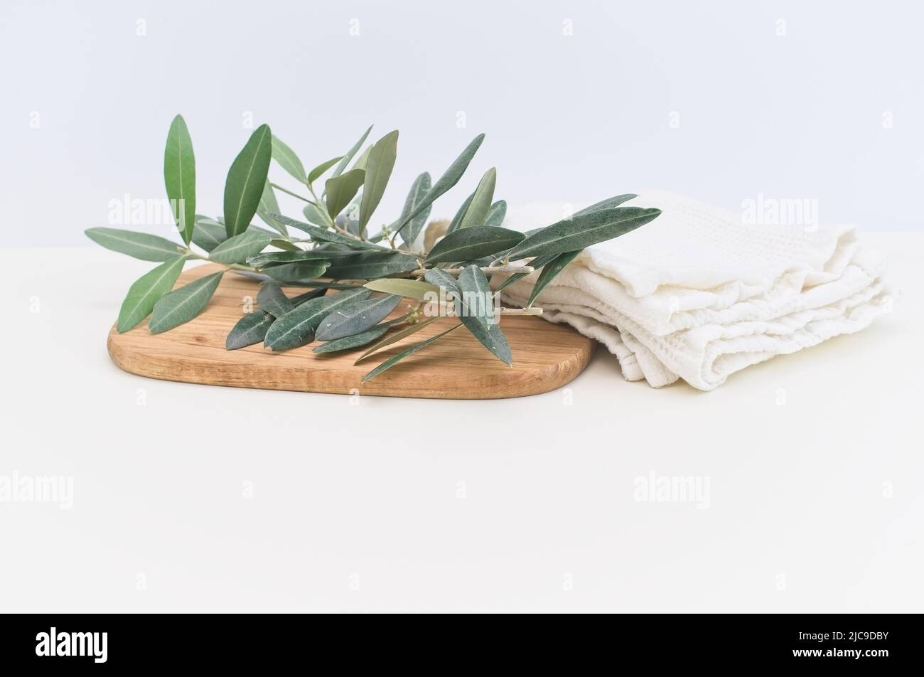 Olivenbaumblätter und Zweige auf einem Holzteller mit weißem Baumwolltuch auf einem hellen Tisch Stockfoto