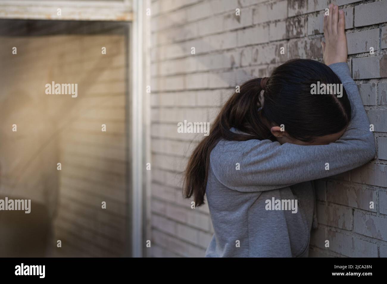 Die junge Frau, die steht, versteckt ihr Gesicht vor einer Backsteinmauer. Probleme von Jugendlichen. Stockfoto