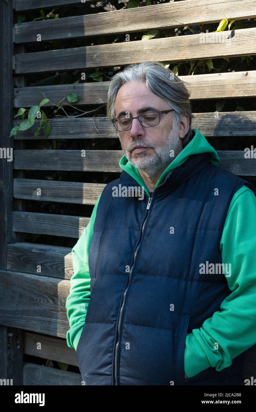 Reifer Mann mit grauem Haar und Brille, der sich auf den Holzzaun lehnt und mit den Händen in den Taschen nachdenklich nach unten blickt. Stockfoto