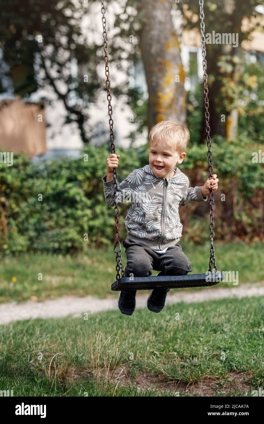 Kleiner Junge, der am sonnigen Sommertag auf dem Spielplatz im öffentlichen Park Spaß auf einer Schaukel hat. Glückliches Kind genießt das Schwingen. Aktive Freizeit im Freien für Kind i Stockfoto