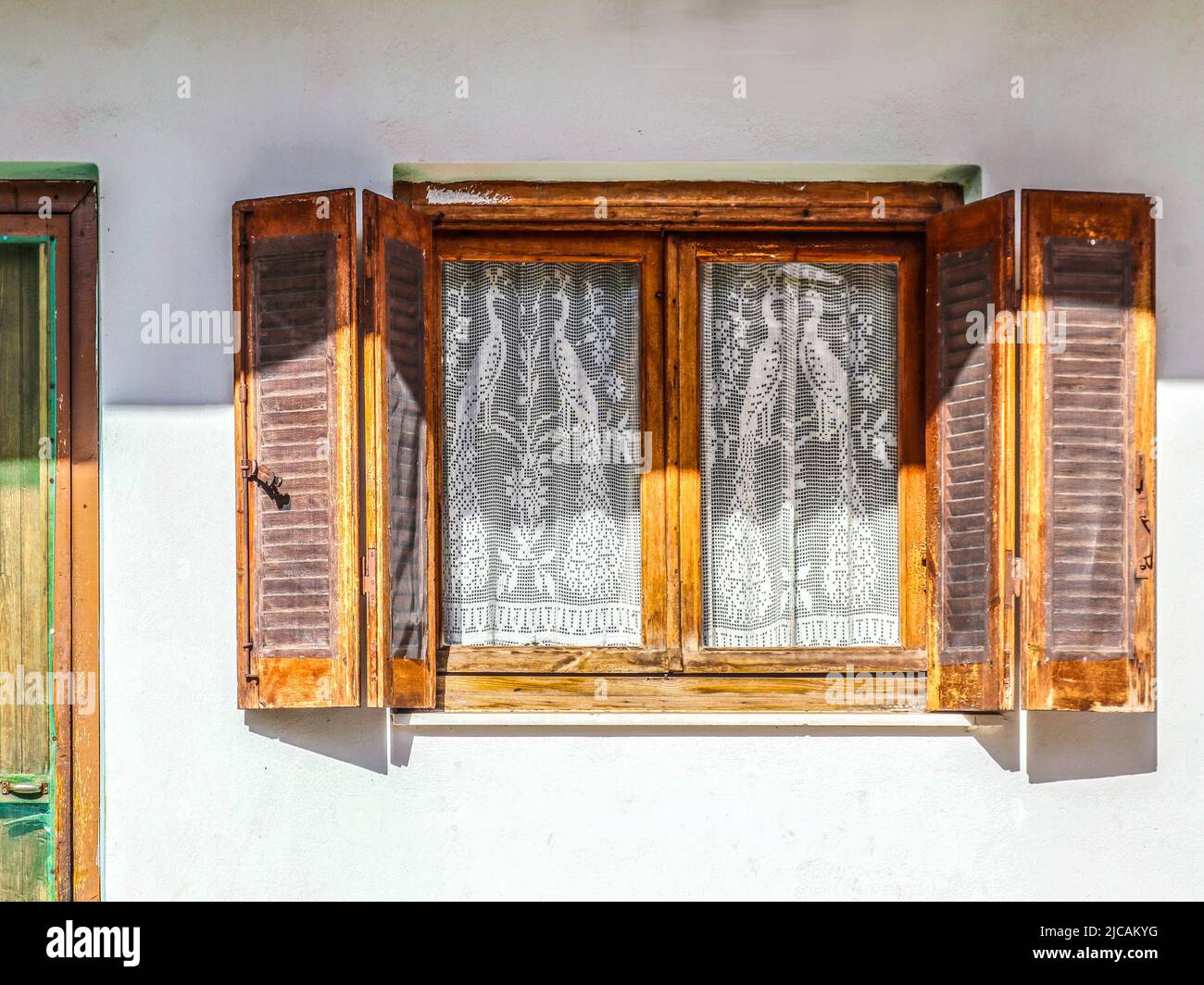 Altes Holzfenster mit offenen Fensterläden an weißer Stuckwand mit aufgehauenen Insektengeflechten und Pfauenvorhängen aus weißer Spitze - Teil der Tür sichtbar - golde Stockfoto