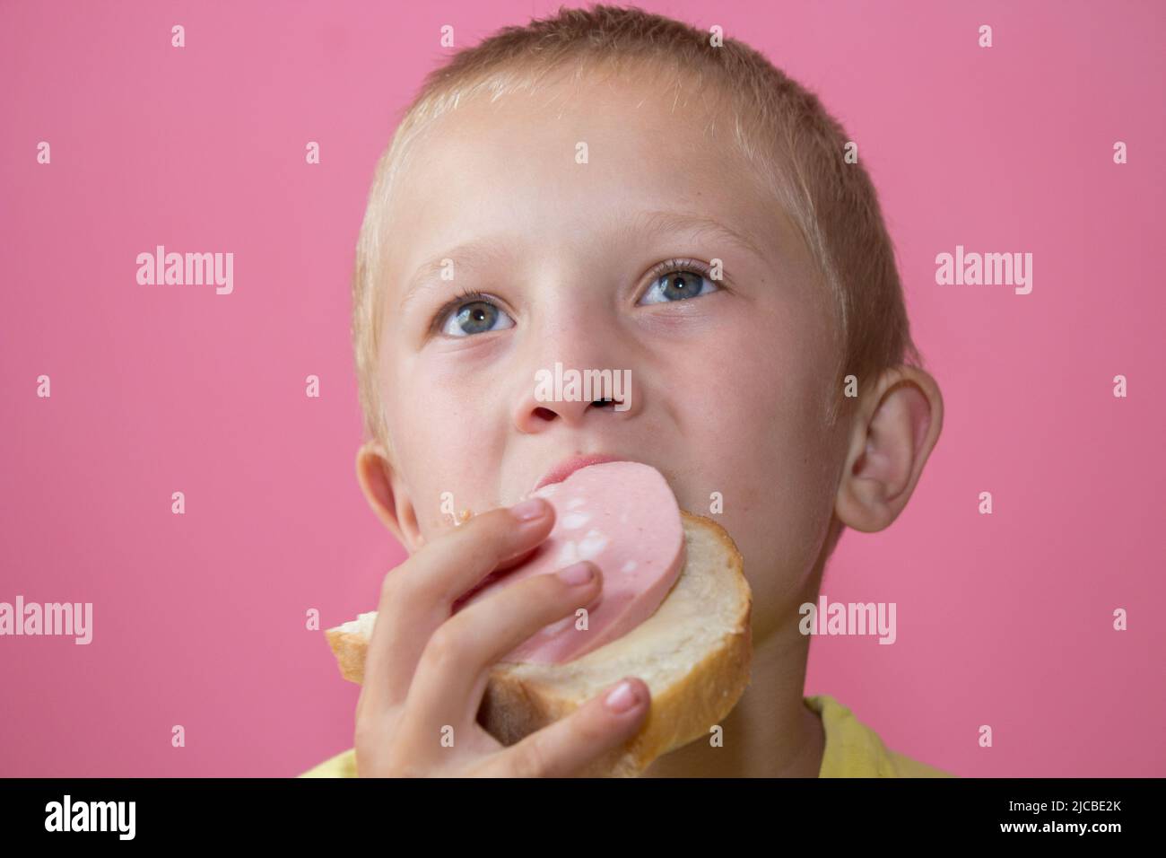 Junge isst ein Sandwich mit gekochter Wurst und sieht interessiert aus Stockfoto