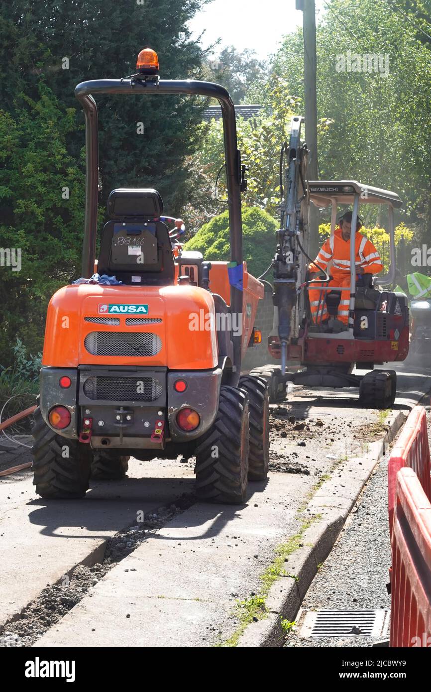 Fiber Optics super schnelle Breitband-Infrastruktur-Projekt in ländlichen Essex Dorf Mini-Bagger Leistungsschalter Fahrer schneidet Bürgersteig Graben Dumper LKW wartet Großbritannien Stockfoto