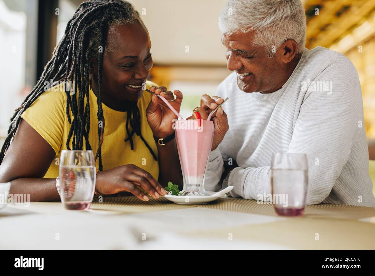 Glückliches reifes Paar lächelt einander an, während sie einen leckeren Milchshake teilen. Fröhliches Seniorenpaar, das sich in einem Restaurant gut Vergnügen hat. Carefree Matte Stockfoto